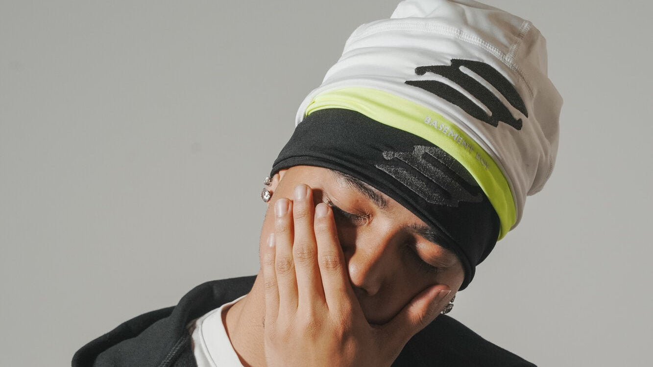 A person wearing a white beanie with black and neon yellow-green stripes, a black headband, a black zip-up hoodie, and a white t-shirt, posing with their face partially covered by their hand against a plain gray background.