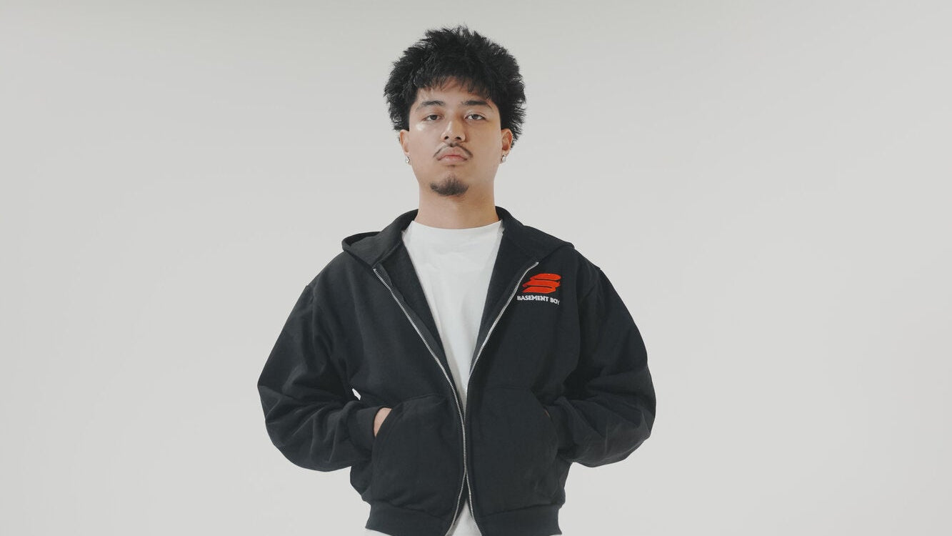 A man modeling a black zip-up hoodie and matching black sweatpants with graphic prints, styled with a white t-shirt, white socks, and brown fuzzy slippers against a plain light gray studio background.